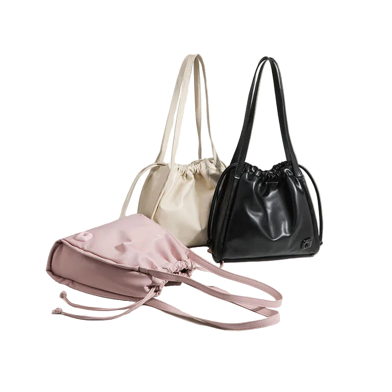 Three drawstring bucket bags in black, cream, and light pink are arranged on a warm beige background. The light pink bag in the foreground is slightly open, showing its interior, while the other two stand closed.