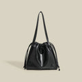 A black faux-leather drawstring bucket bag with a single shoulder strap, shown against a solid beige background with a small logo on the bottom right corner.
